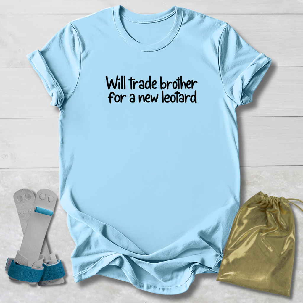 A light blue graphic t-shirt with the text 'Will trade brother for a new leopard' printed in black on the chest, displayed in a flat-lay arrangement with a gold drawstring bag and a pair of gray and blue athletic shoes on a white wood background.