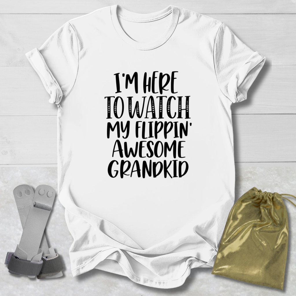 A white short-sleeve graphic T-shirt laid flat on a light wood background, with black text reading 'I'M HERE TO WATCH MY FLIPPIN' AWESOME GRANDKID.' Gray flip-flops and a small metallic gold drawstring pouch are placed beside it as styling props.