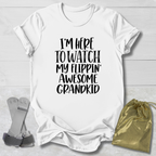 A white short-sleeve graphic T-shirt laid flat on a light wood background, with black text reading 'I'M HERE TO WATCH MY FLIPPIN' AWESOME GRANDKID.' Gray flip-flops and a small metallic gold drawstring pouch are placed beside it as styling props.