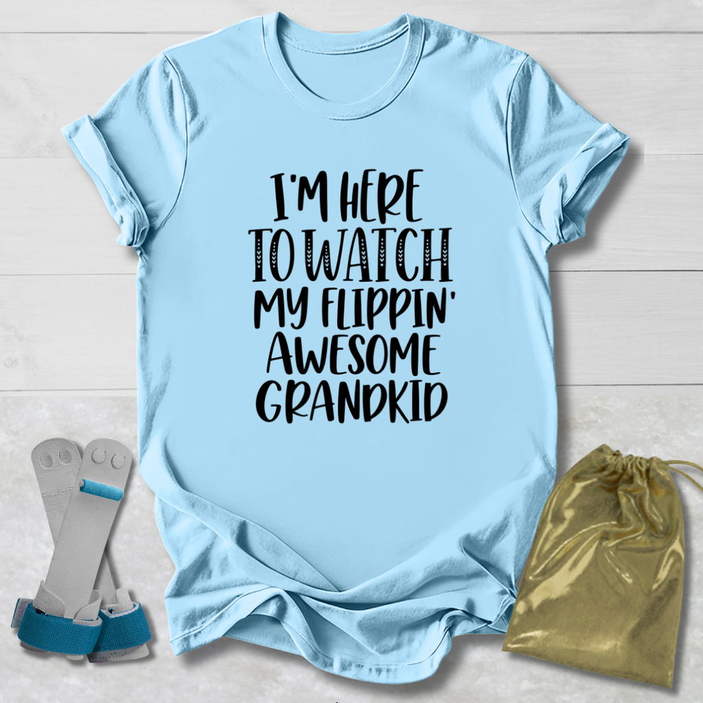A light blue graphic t-shirt laid flat on a white wood background, featuring the text 'I'M HERE TO WATCH MY FLIPPIN' AWESOME GRANDKID' in black. A pair of gray and blue baby support braces and a gold drawstring pouch are styled alongside the shirt.