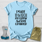 A light blue graphic t-shirt laid flat on a white wood background, featuring the text 'I'M HERE TO WATCH MY FLIPPIN' AWESOME GRANDKID' in black. A pair of gray and blue baby support braces and a gold drawstring pouch are styled alongside the shirt.