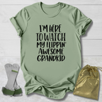 A sage green graphic t-shirt with black text reading 'I'M HERE TO WATCH MY FLIPPIN' AWESOME GRANDKID' is displayed in a flat-lay arrangement on a white wood background, accompanied by a pair of white and green supportive leg braces on the left and a shiny gold drawstring bag on the right.