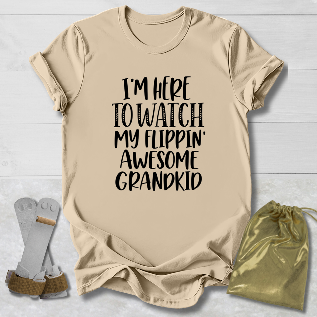 A beige graphic t-shirt with the text 'I'M HERE TO WATCH MY FLIPPIN' AWESOME GRANDKID' in black. The t-shirt is displayed in a flat-lay arrangement on a white wood background, accompanied by a pair of white and brown flip-flops on the left and a shiny gold drawstring pouch on the right.