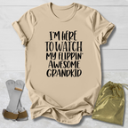 A beige graphic t-shirt with the text 'I'M HERE TO WATCH MY FLIPPIN' AWESOME GRANDKID' in black. The t-shirt is displayed in a flat-lay arrangement on a white wood background, accompanied by a pair of white and brown flip-flops on the left and a shiny gold drawstring pouch on the right.