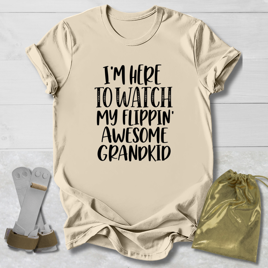 A beige graphic t-shirt with black text reading 'I'M HERE TO WATCH MY FLIPPIN' AWESOME GRANDKID' is displayed in a flat-lay arrangement on a white wood background, accompanied by a pair of flip-flops and a gold drawstring pouch.