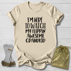 A beige graphic t-shirt with black text reading 'I'M HERE TO WATCH MY FLIPPIN' AWESOME GRANDKID' is displayed in a flat-lay arrangement on a white wood background, accompanied by a pair of flip-flops and a gold drawstring pouch.
