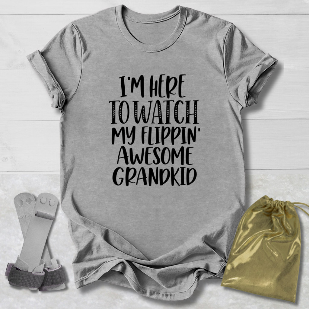 A heather gray graphic t-shirt with black text reading 'I'M HERE TO WATCH MY FLIPPIN' AWESOME GRANDKID' is laid flat on a white wood background, accompanied by a gold drawstring pouch and a pair of silver-and-gray sandals.