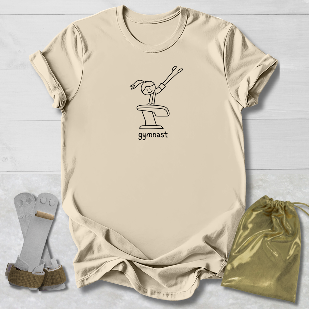 A beige short-sleeve T-shirt laid flat on a light wood background, featuring a gymnast on a balance beam graphic with the word 'GYMNAST' underneath. A shiny gold drawstring bag and a pair of gymnastics shoes are placed beside the shirt as styling props.