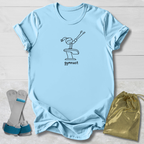 A light blue short-sleeve T-shirt laid flat on a white wood background, printed with a simple black line drawing of a gymnast and the word 'gymnast' below. Two gray and blue gymnastics bars are placed to the lower left, and a shiny metallic gold drawstring bag is to the lower right.