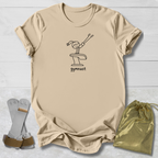 A beige graphic t-shirt featuring a black line art illustration of a gymnast on a vaulting table with the word 'gymnast' underneath. The t-shirt is displayed in a flat-lay arrangement alongside a pair of gymnast straps and a shiny gold drawstring bag on a white wood background.
