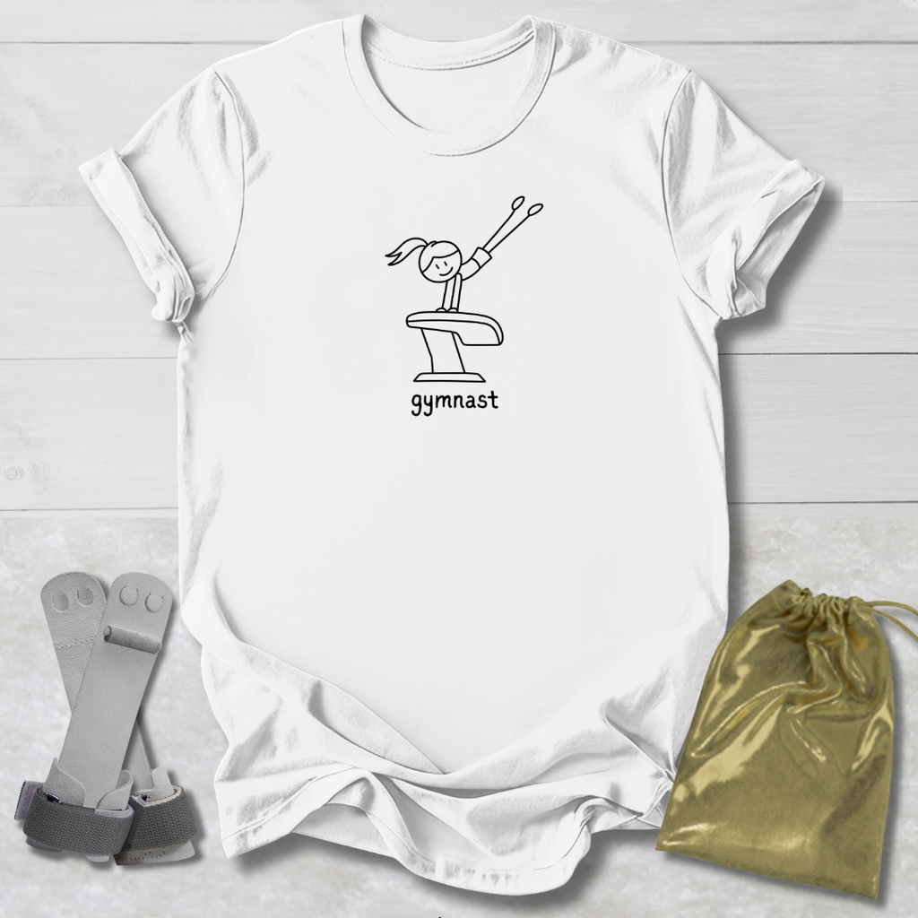 A white short-sleeve T-shirt laid flat on a light wood background, featuring a black line drawing of a gymnast on a vaulting table with the word 'gymnast' underneath. A gold drawstring pouch and gray gymnast straps are placed beside the shirt as styling props.