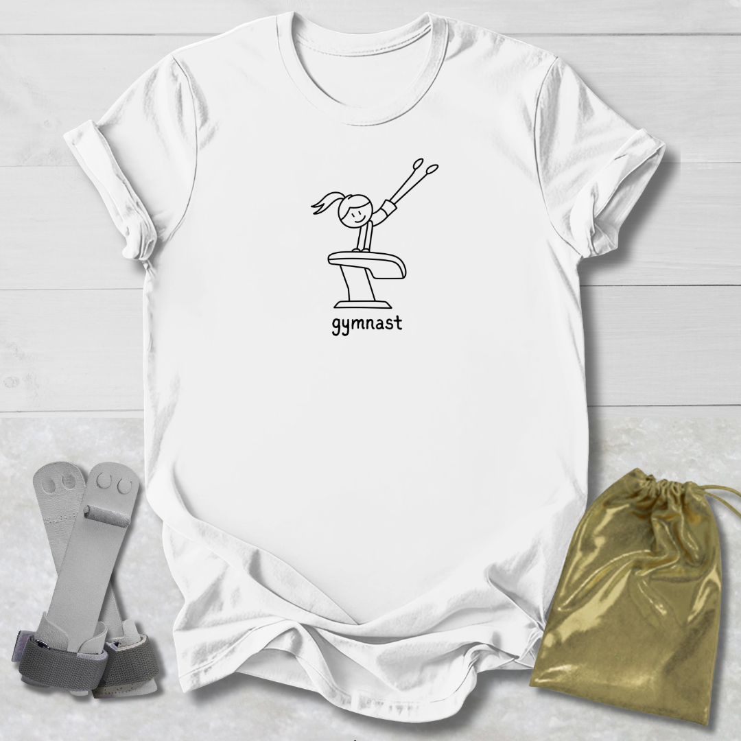 A white short-sleeve T-shirt laid flat on a light wood background, featuring a black line drawing of a gymnast on a vaulting table with the word 'gymnast' underneath. A gold drawstring pouch and gray gymnast straps are placed beside the shirt as styling props.