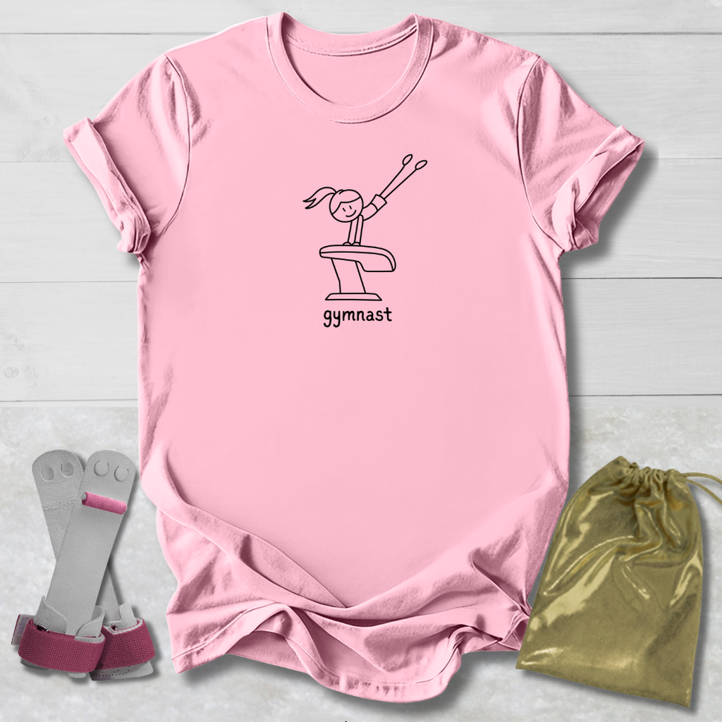 A light pink short-sleeve t-shirt with a black line art graphic of a gymnast on a vaulting horse and the word 'gymnast' printed below. The t-shirt is laid flat on a light gray wood background, accompanied by a pair of pink and gray gymnastics straps on the left and a shiny gold drawstring bag on the right.