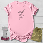 A light pink short-sleeve t-shirt with a black line art graphic of a gymnast on a vaulting horse and the word 'gymnast' printed below. The t-shirt is laid flat on a light gray wood background, accompanied by a pair of pink and gray gymnastics straps on the left and a shiny gold drawstring bag on the right.