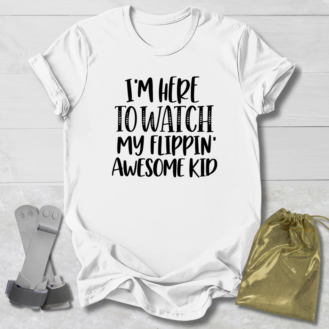 A white short-sleeve t-shirt with black text reading 'I'M HERE TO WATCH MY FLIPPIN' AWESOME KID', styled in a flat-lay with a gold drawstring bag and a pair of gray flip-flops on a light wood background.