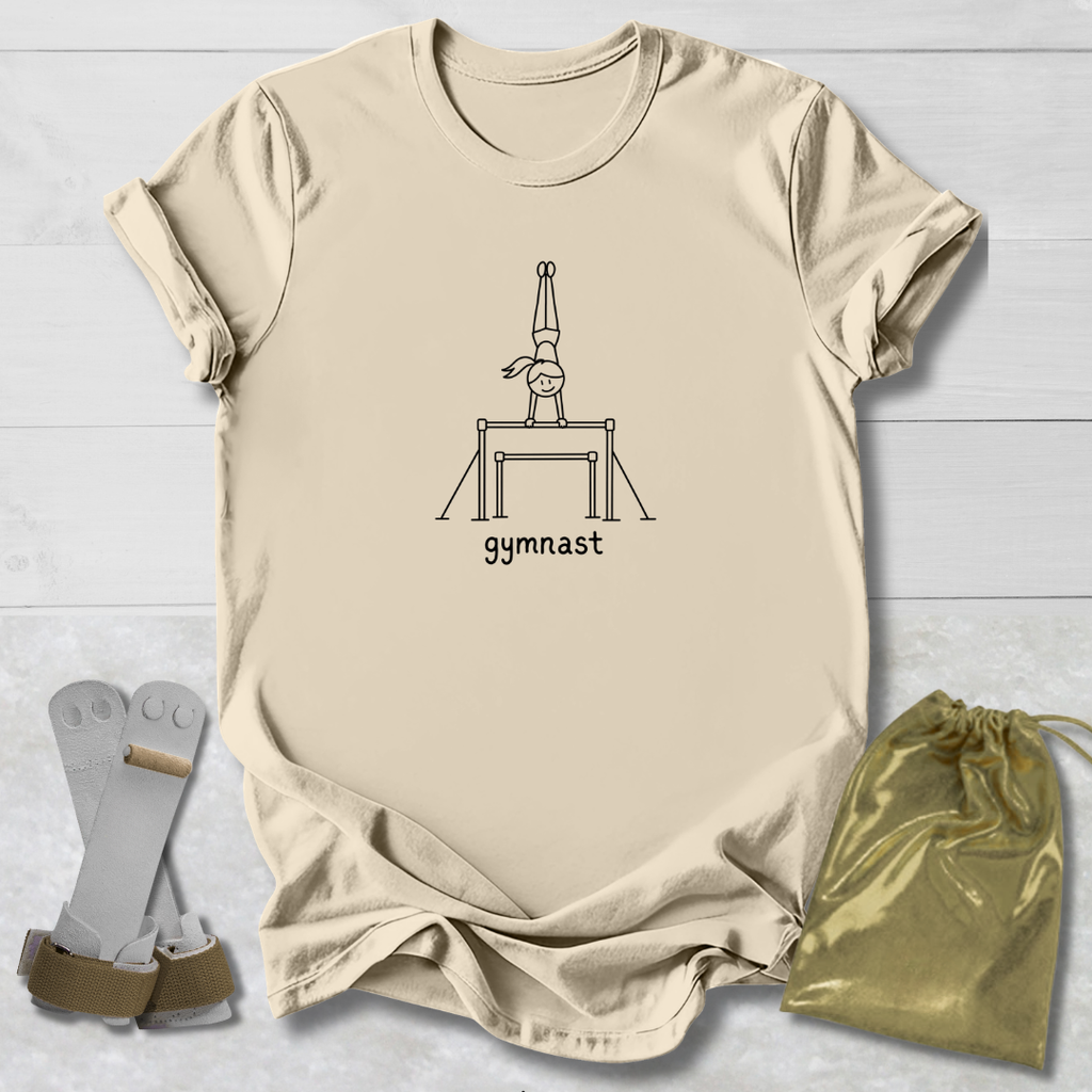 A beige short-sleeve T-shirt laid flat on a white wood background, featuring a black line drawing of a gymnast on a balance bar with the word 'gymnast' underneath. A pair of gymnastics straps and a shiny gold drawstring bag are placed beside the shirt as styling props.