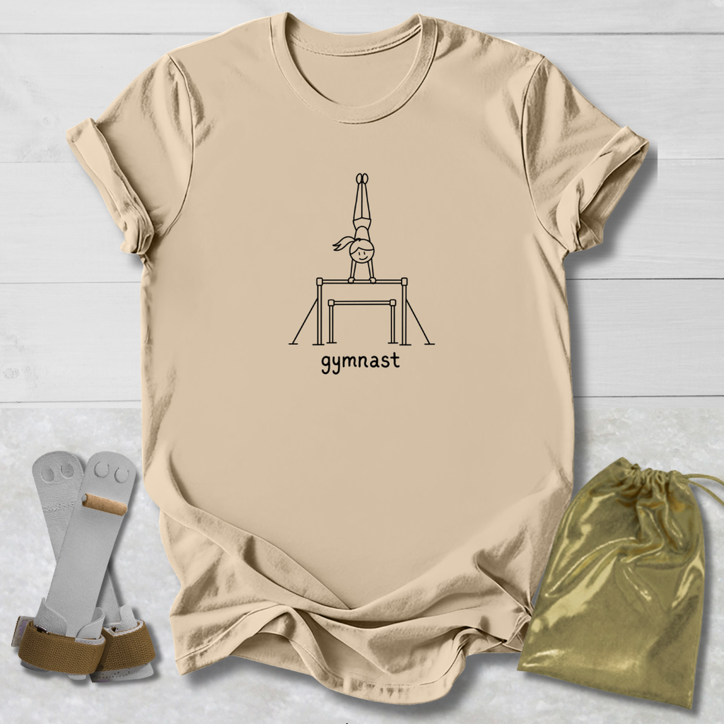 A beige graphic t-shirt featuring a black line art illustration of a gymnast on a balance beam with the word 'gymnast' printed below. The t-shirt is laid flat on a white wood background alongside a pair of chalk gloves and a shiny gold drawstring bag.