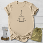A beige graphic t-shirt featuring a black line art illustration of a gymnast on a balance beam with the word 'gymnast' printed below. The t-shirt is laid flat on a white wood background alongside a pair of chalk gloves and a shiny gold drawstring bag.