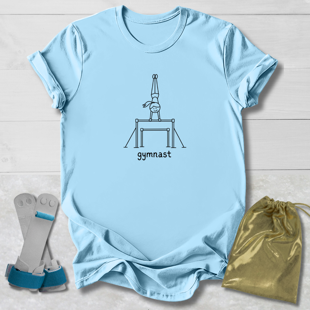 A light blue short-sleeve T-shirt laid flat on a white wood background, featuring a black line drawing of a gymnast on a bar and the word 'gymnast' printed below. A pair of gray and blue gymnastic straps and a shiny gold drawstring bag are placed beside the shirt as styling props.