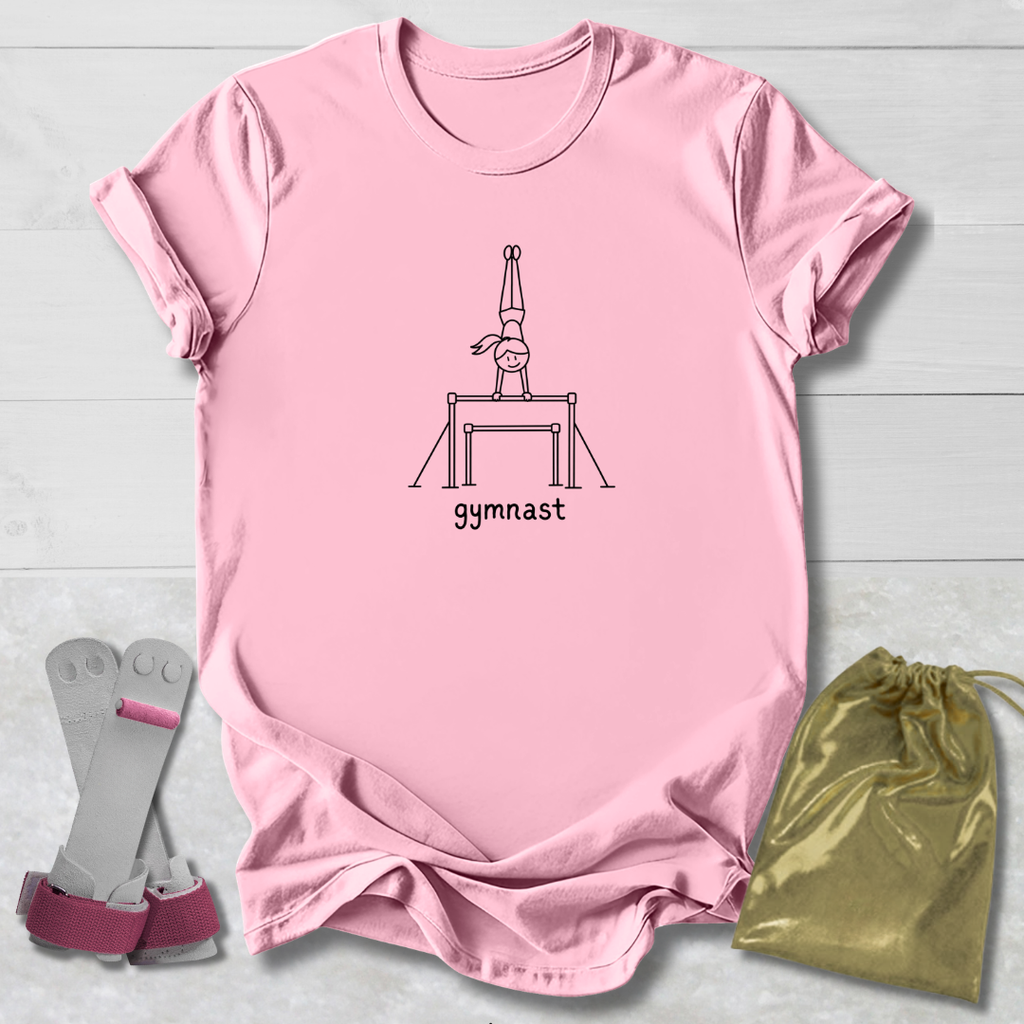 A light pink short-sleeve t-shirt laid flat on a white wood background, featuring a black line art graphic of a gymnast on a bar and the word 'gymnast' printed below. A pair of pink and gray gymnastics straps and a shiny gold drawstring bag are placed beside the shirt as styling props.