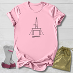 A light pink short-sleeve t-shirt laid flat on a white wood background, featuring a black line art graphic of a gymnast on a bar and the word 'gymnast' printed below. A pair of pink and gray gymnastics straps and a shiny gold drawstring bag are placed beside the shirt as styling props.