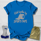 A bright blue graphic T-shirt laid flat on a white wood background, printed with the phrase 'Held together by SPORTS TAPE' and a roll of sports tape graphic. Two white and blue athletic wrist wraps are placed beside the shirt, along with a shiny gold drawstring pouch.