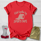 A red short-sleeve T-shirt laid flat on a white wood background, printed with the phrase 'Held together by SPORTS TAPE' and a roll of sports tape graphic. A pair of white and red athletic wrist wraps and a shiny gold drawstring pouch are placed beside the shirt as included accessories or related items.