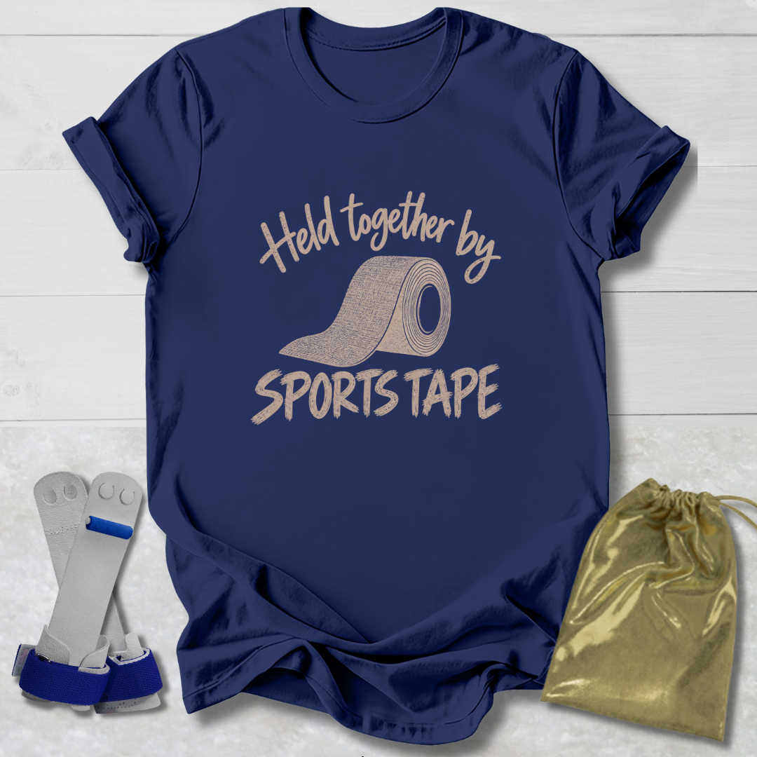 A navy blue graphic T-shirt laid flat on a white wood background, printed with the phrase 'Held together by SPORTS TAPE' and a roll of sports tape illustration. A pair of white and blue athletic tape rolls and a shiny gold drawstring pouch are placed beside the shirt as included accessories.