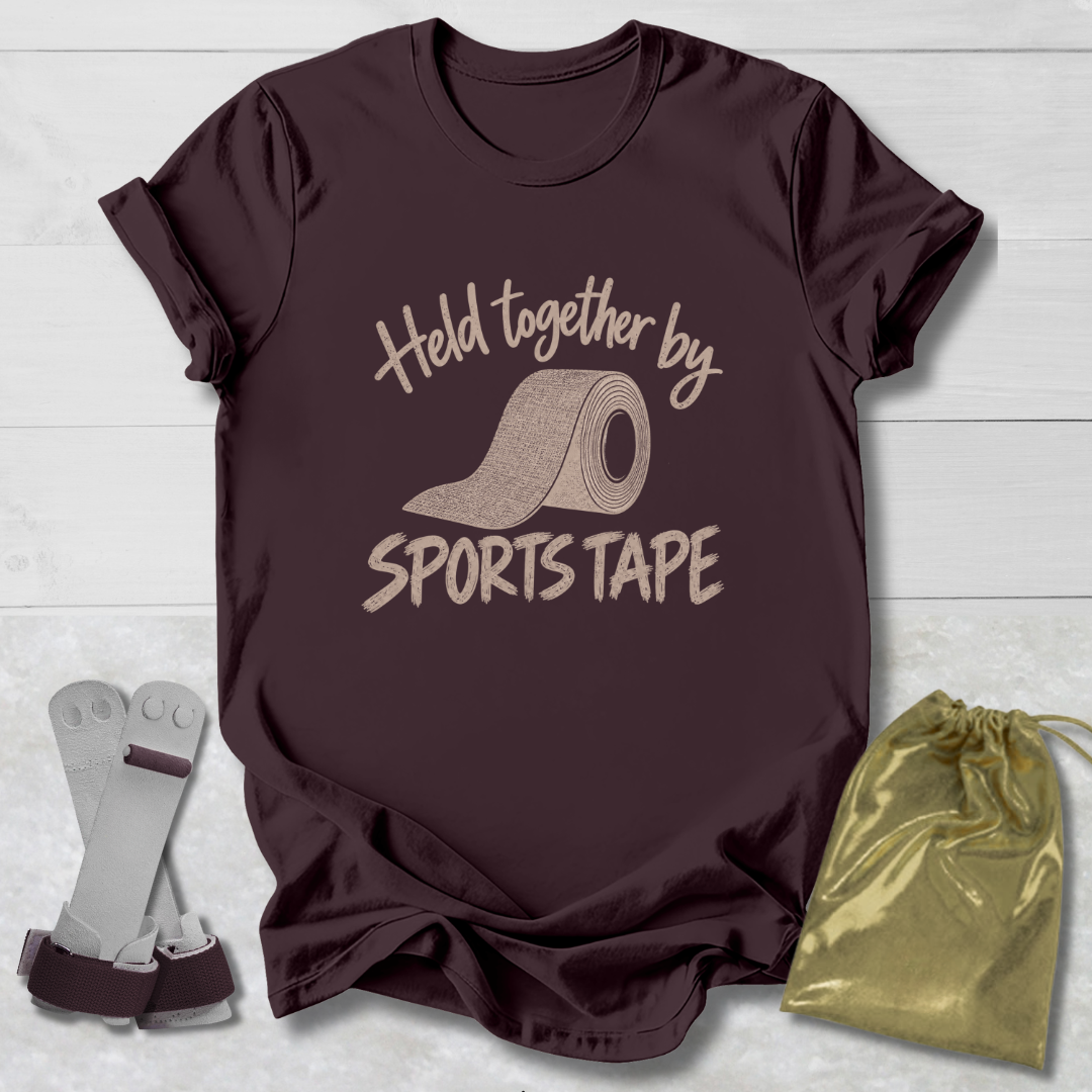 A dark brown short-sleeve t-shirt with a beige graphic that reads 'Held together by SPORTS TAPE' around an illustration of a roll of sports tape. The shirt is displayed on a light gray wood background alongside a pair of gray wrist wraps and a shiny gold drawstring pouch.