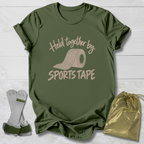 An olive green graphic t-shirt laid flat on a white wood background, featuring a beige print that reads 'Held together by SPORTS TAPE' with a roll of sports tape illustration. A pair of white and green athletic wrist wraps and a shiny gold drawstring pouch are arranged around the shirt.