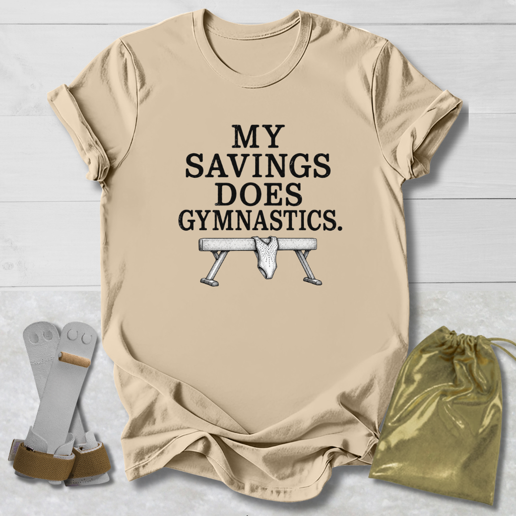 A beige graphic t-shirt laid flat on a white wood background, featuring the text 'MY SAVINGS DOES GYMNASTICS.' above a bench press illustration.