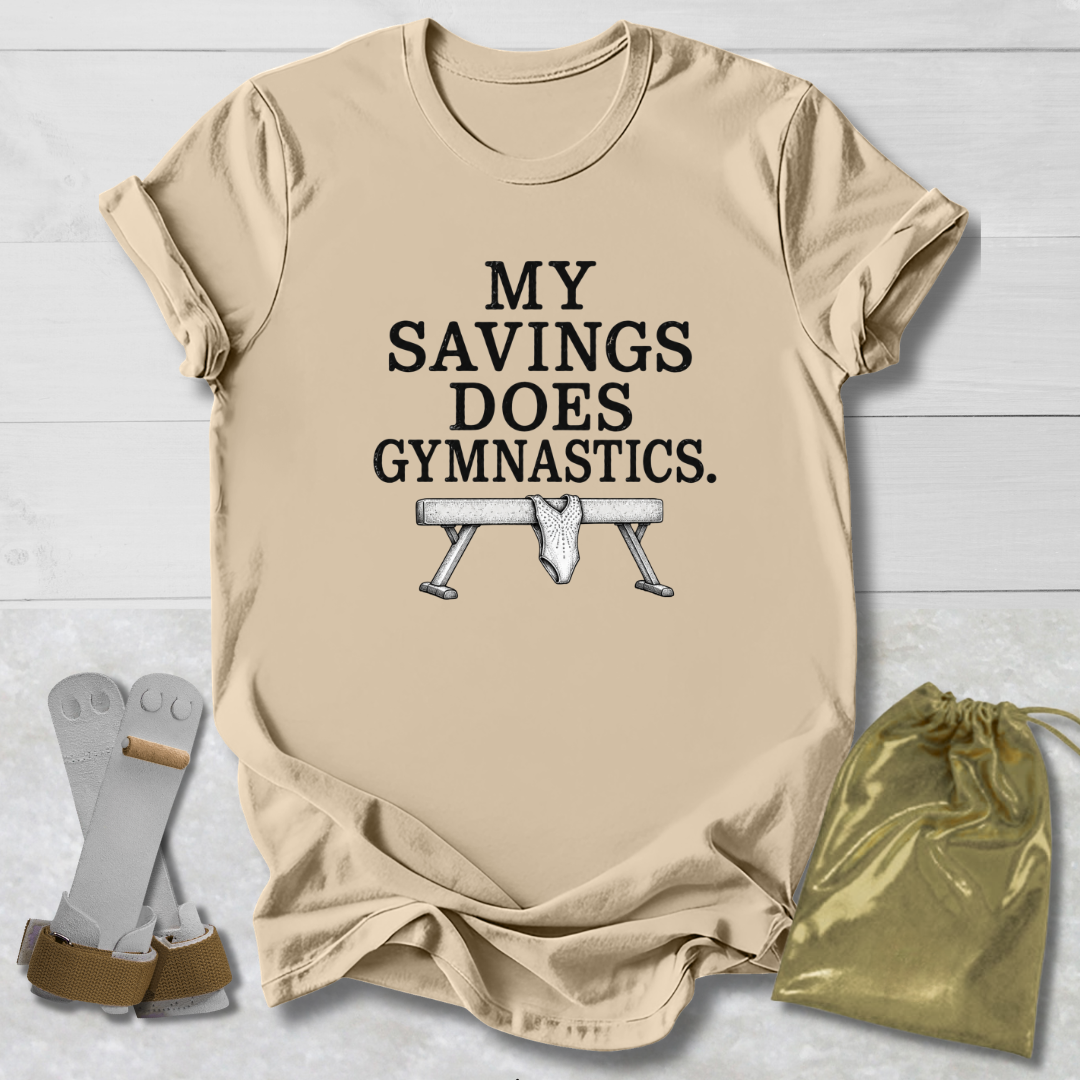 A beige graphic t-shirt laid flat on a white wood background, featuring the text 'MY SAVINGS DOES GYMNASTICS.' above a bench press illustration.