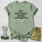 A sage green graphic t-shirt laid flat on a white wood background, featuring the text 'MY SAVINGS DOES GYMNASTICS.' and a bench press illustration.