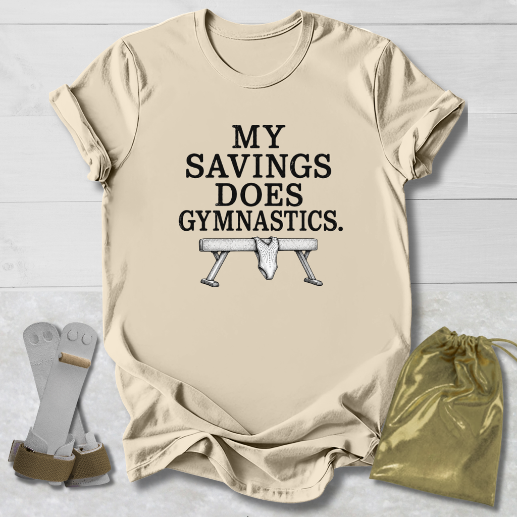 A beige graphic t-shirt laid flat on a white wood background, featuring the text 'MY SAVINGS DOES GYMNASTICS.' above a bench press illustration.