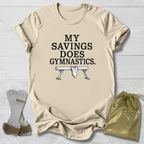 A beige graphic t-shirt laid flat on a white wood background, featuring the text 'MY SAVINGS DOES GYMNASTICS.' above a bench press illustration.