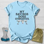 A light blue graphic T-shirt laid flat on a white wood background, with the text 'MY SAVINGS DOES GYMNASTICS.' and a bench press illustration.