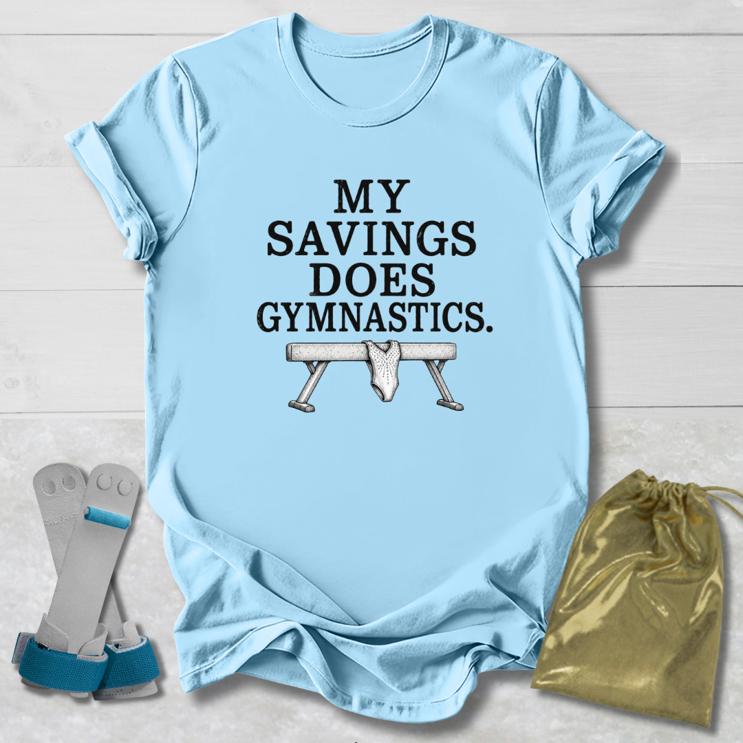 A light blue graphic T-shirt laid flat on a white wood background, with the text 'MY SAVINGS DOES GYMNASTICS.' and a bench press illustration.