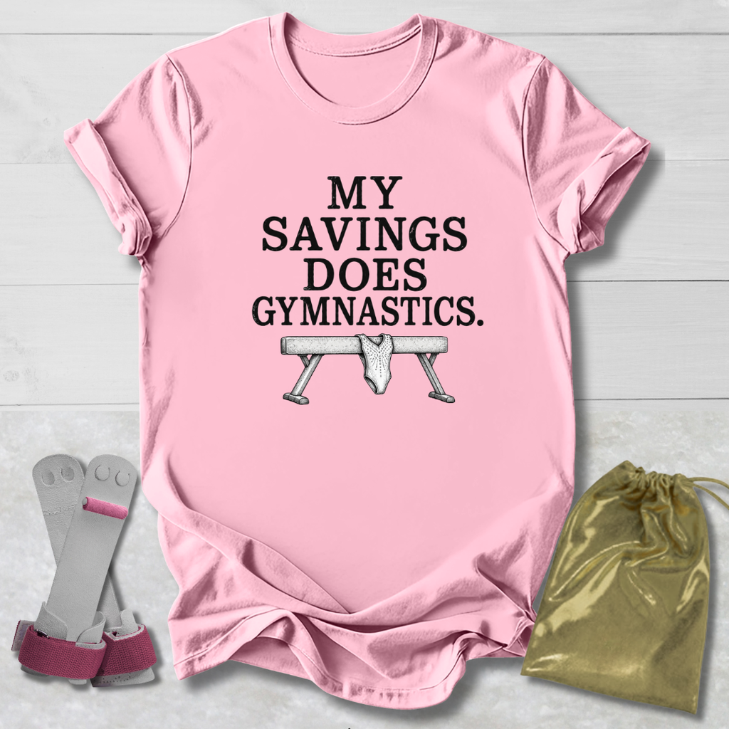 A pink short-sleeve graphic T-shirt laid flat on a light wood background, with the text 'MY SAVINGS DOES GYMNASTICS.' and a bench press illustration printed on the front.