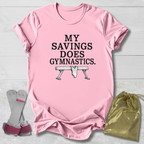 A pink short-sleeve graphic T-shirt laid flat on a light wood background, with the text 'MY SAVINGS DOES GYMNASTICS.' and a bench press illustration printed on the front.