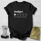 A black graphic t-shirt with white text reading 'Judges' and a one-star rating graphic, styled in a flat lay with a gold drawstring bag and a pair of gray and black wrist/ankle wraps on a light wood background.