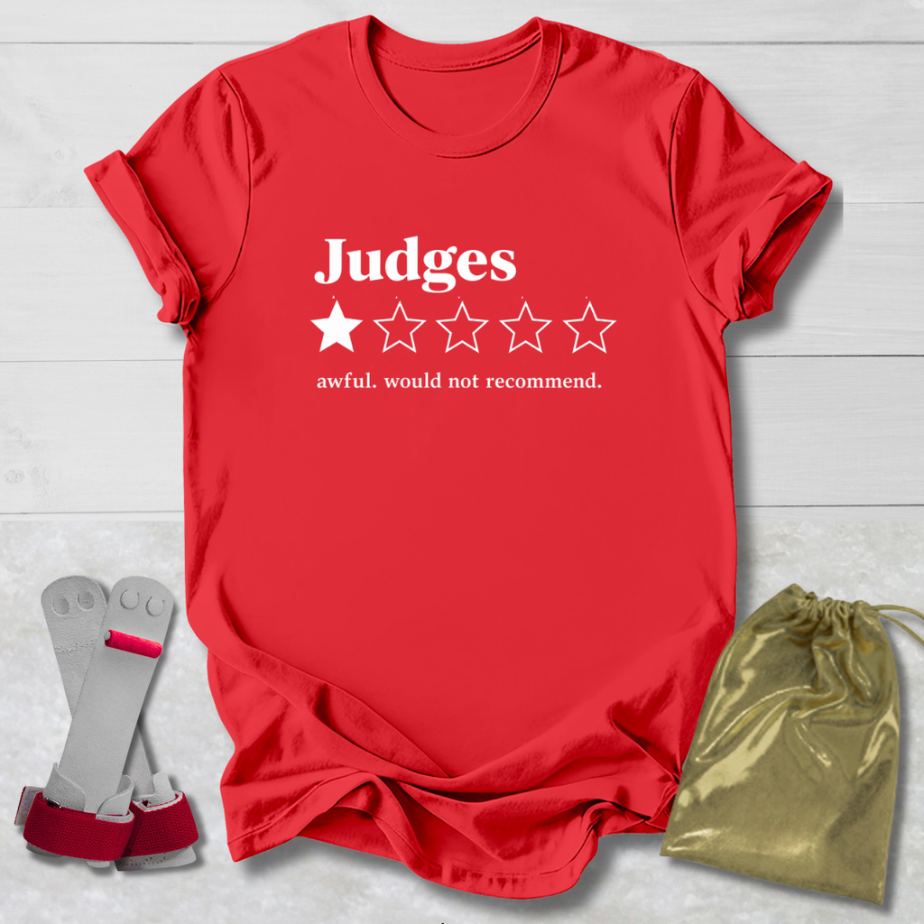 A red short-sleeve t-shirt with white text reading 'Judges' and a one-star rating graphic, styled on a light wood background with a gray-and-red wrist strap and a gold drawstring pouch.