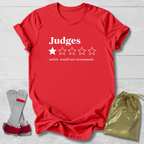 A red short-sleeve t-shirt with white text reading 'Judges' and a one-star rating graphic, styled on a light wood background with a gray-and-red wrist strap and a gold drawstring pouch.