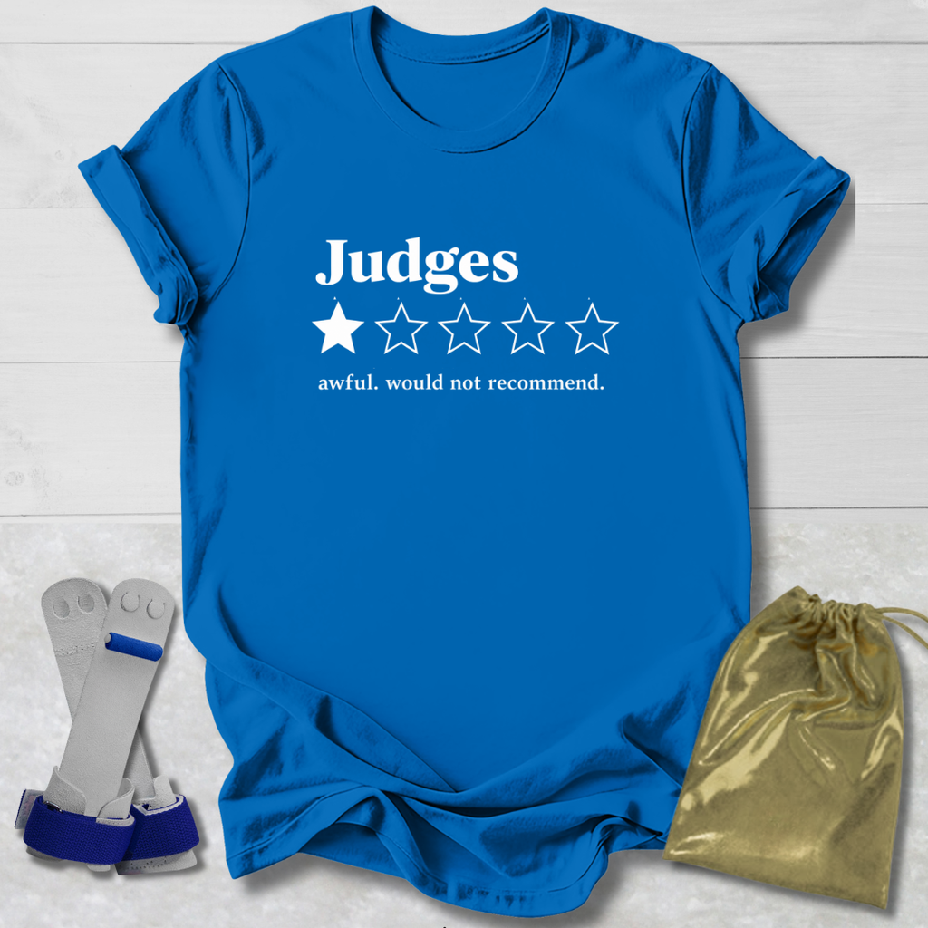A bright blue graphic T-shirt laid flat on a white wood background, with white text reading 'Judges' and a one-star rating graphic. Two gray-and-blue wrist restraints and a shiny gold drawstring pouch are placed beside the shirt as included accessories.
