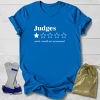 A bright blue graphic T-shirt laid flat on a white wood background, with white text reading 'Judges' and a one-star rating graphic. Two gray-and-blue wrist restraints and a shiny gold drawstring pouch are placed beside the shirt as included accessories.
