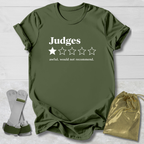 An olive green short-sleeve t-shirt laid flat on a white wood background, printed with the word 'Judges' and a one-star rating graphic. Two small accessories are placed beside it: a pair of white and green wrist/ankle wraps and a shiny gold drawstring pouch.