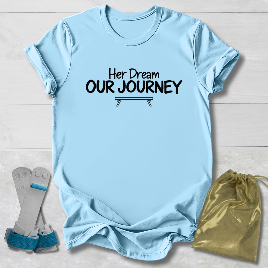 A light blue graphic t-shirt with the text 'Her Dream OUR JOURNEY' printed in black on the chest, styled in a flat-lay with a pair of gray and blue leg wraps and a gold drawstring bag on a white wood background.