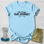A light blue graphic t-shirt with the text 'Her Dream OUR JOURNEY' printed in black on the chest, styled in a flat-lay with a pair of gray and blue leg wraps and a gold drawstring bag on a white wood background.