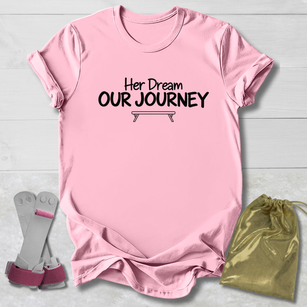 A light pink short-sleeve t-shirt with the black printed text 'Her Dream OUR JOURNEY' and a small bench graphic, laid flat on a white wood background. A pair of pink and gray leg wraps and a gold drawstring bag are placed beside it as styling props.