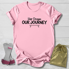 A light pink short-sleeve t-shirt with the black printed text 'Her Dream OUR JOURNEY' and a small bench graphic, laid flat on a white wood background. A pair of pink and gray leg wraps and a gold drawstring bag are placed beside it as styling props.