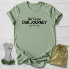 A sage green graphic t-shirt with the black printed text 'Her Dream OUR JOURNEY' and a small bench graphic, displayed on a white wood background alongside a pair of white and green support braces and a gold drawstring bag.